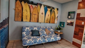 Een kamer met een blauwe gestreepte bank, een muur versierd met surfplanken, een klein houten bijzettafeltje met een plant en een houten deur.