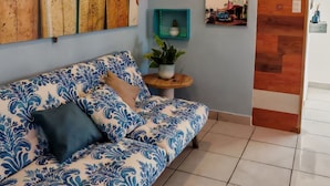 Een woonkamer met een gestreept sofa, een surfboard aan de muur, een kleine tafel met een plant en een houten deur.