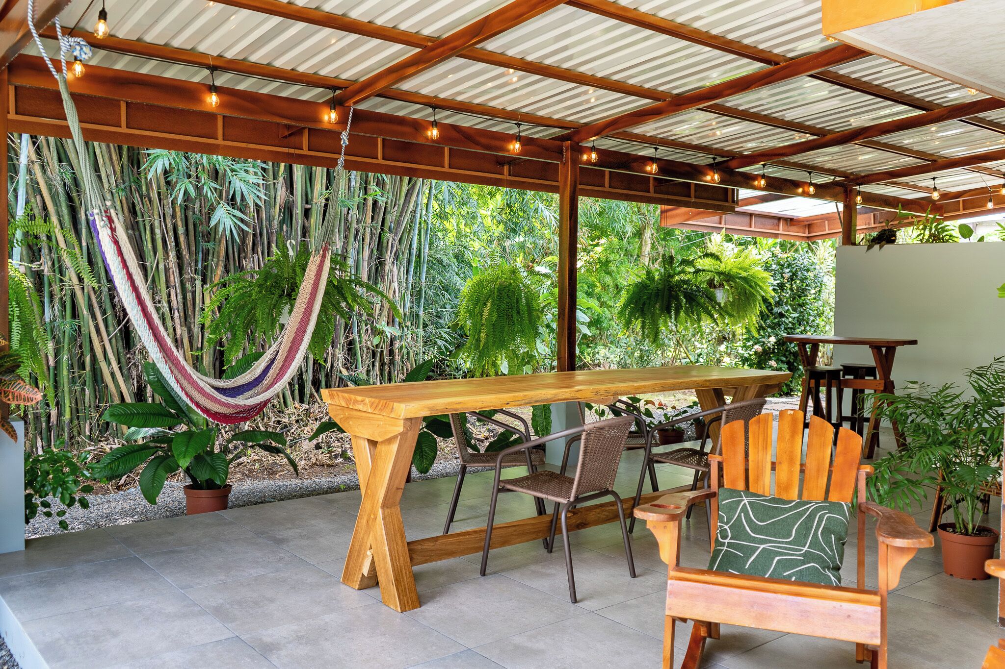 A covered outdoor seating area with a wooden table and chairs, surrounded by lush greenery and a hammock.