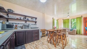 A kitchen with a microwave, toaster, and coffee maker, a dining area with a table and chairs, and a living room with a sofa and curtains. - Hummingbird jungle (La Fortuna)