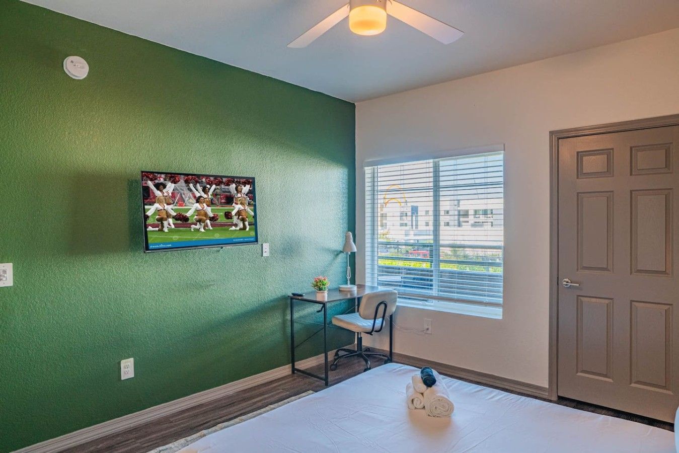 A room with a green wall, a television mounted on it, a window with blinds, a door, and a bed with a rolled-up towel on it.