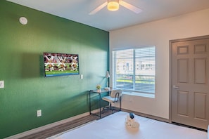 A hotel room with a green wall, a bed with a rolled-up towel, a desk with a chair, a window with blinds, and a door.