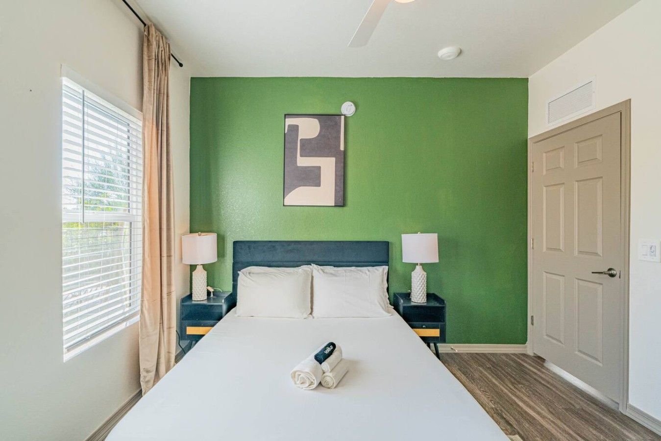 A hotel room with a bed, two bedside tables with lamps, a green accent wall, a door, and a window with blinds.