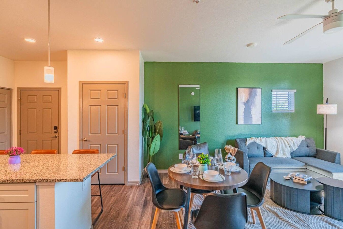 A modern living room with a green accent wall, a dining area with a round table and chairs, and a kitchen with a granite countertop.