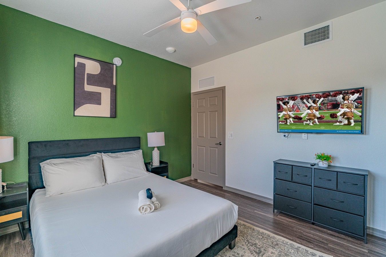 A hotel room with a bed, a ceiling fan, a television, and a door.