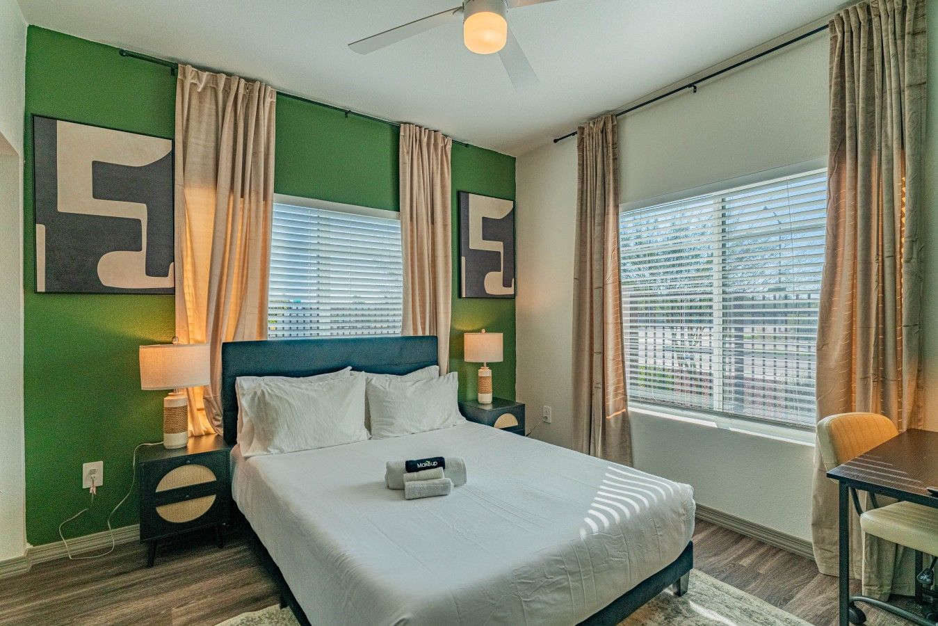 A hotel room with a bed, two bedside tables, a desk, and a window with blinds.