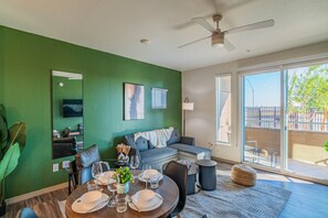 A modern living room with a green accent wall, a ceiling fan, a sofa, a dining table set for four, and a large window offering a view of the outdoors. - Walk To Stadium with Balcony-Pool-Gym-Parking-W/D (Glendale)