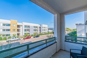 A balcony with a view of a multi-story building, a street with parked cars, and a clear sky.