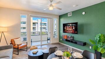 Uma sala de estar moderna com TV de tela plana, espaço de jantar para seis pessoas e uma varanda com vista para a paisagem urbana.