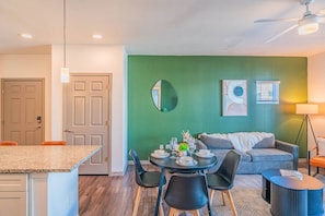 A modern living room with a green accent wall, a round mirror, and a dining area with a granite countertop and black chairs.