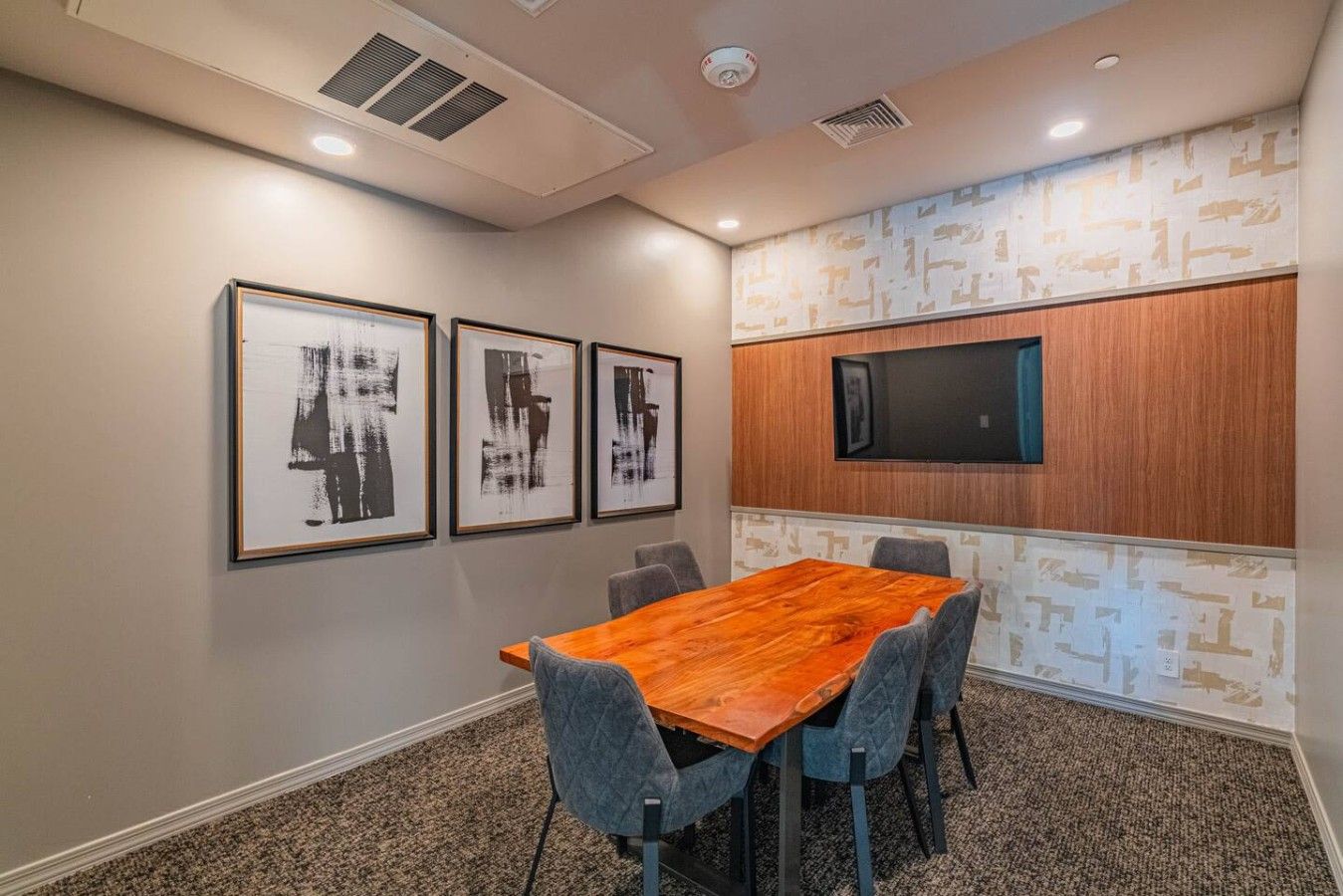 A conference room with a wooden table, six chairs, and three framed abstract paintings on the wall.