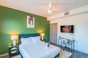 A hotel room with a bed, two bedside lamps, a ceiling fan, a TV on the wall, and a desk with a chair.