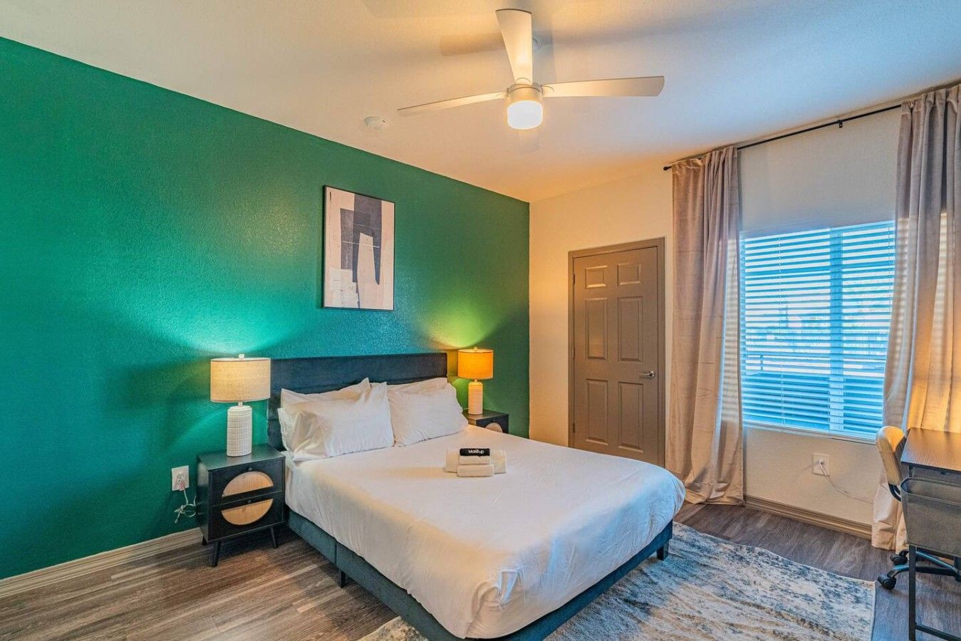 A hotel room with a bed, two bedside tables, a ceiling fan, a door, and a window with curtains.