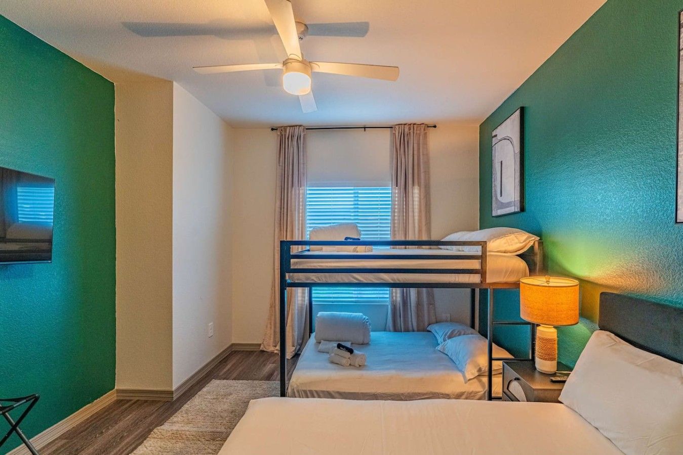 Ein Zimmer mit Etagenbetten, einem Deckenventilator, einem Fenster mit Vorhängen, einer Nachttischlampe und einem an der Wand montierten Fernseher.