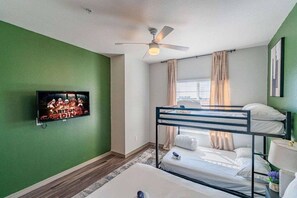 A room with a bunk bed, a ceiling fan, a television mounted on the wall, and a window with curtains.