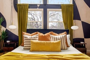 Ein Bett mit vielen Kissen, einem Nachttisch, einer Lampe und einem Fenster mit Vorhängen.