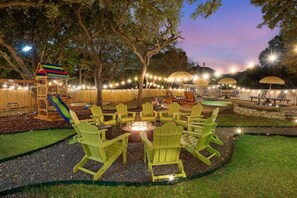 Un jardin avec un foyer, des chaises vertes, un toboggan et des guirlandes lumineuses.