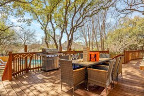 Terraza exterior con parrilla, mesa de comedor y sillas, con vista a una piscina y árboles.
