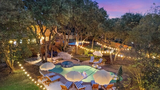 An outdoor swimming pool area with lounge chairs and umbrellas, surrounded by trees and string lights. - HeatedPool PickleBall Basktball Golf FirePit Games (San Antonio)