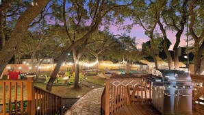 A wooden deck with a barbecue grill, surrounded by trees and string lights. - HeatedPool PickleBall Basktball Golf FirePit Games (San Antonio)