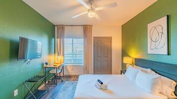 A hotel room with a bed, a ceiling fan, a TV, a desk with a lamp, and a window with curtains.