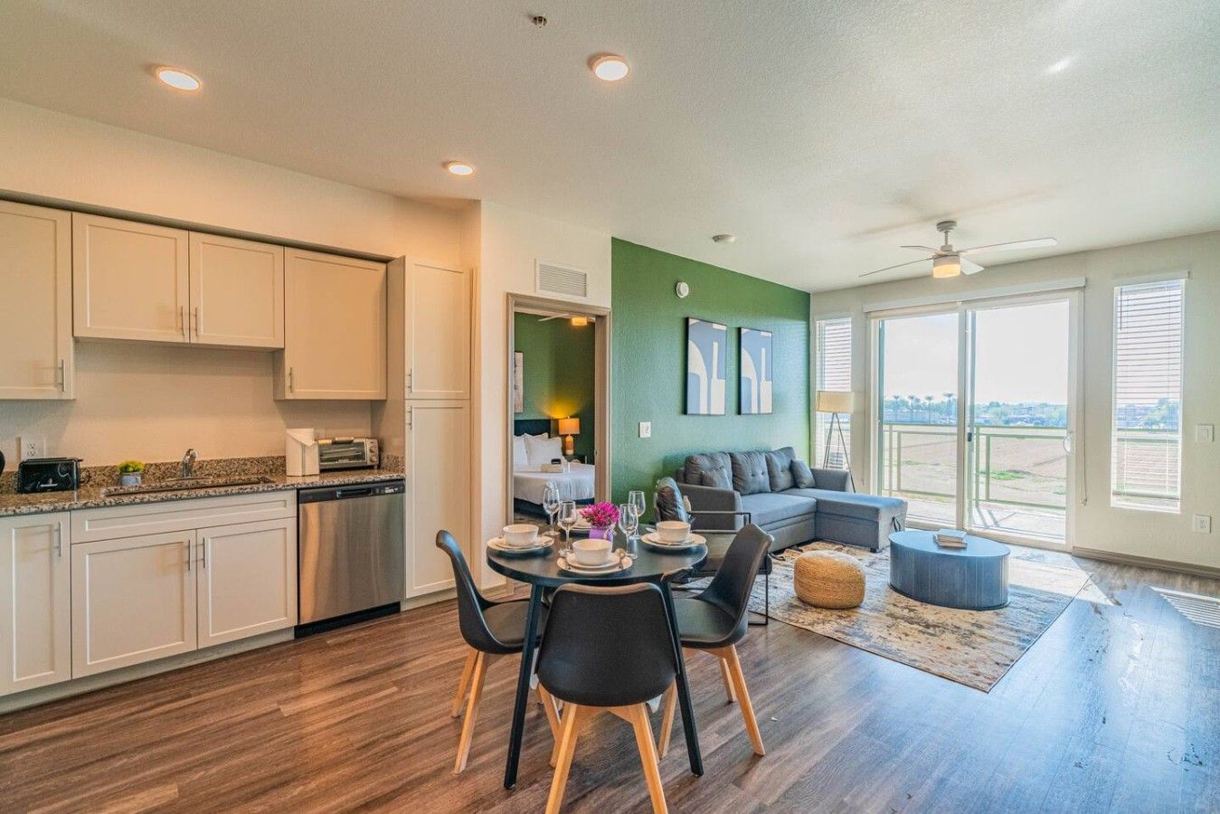 A modern kitchen with white cabinets, a dining area with a round table and chairs, a gray sofa, and a balcony with a view.