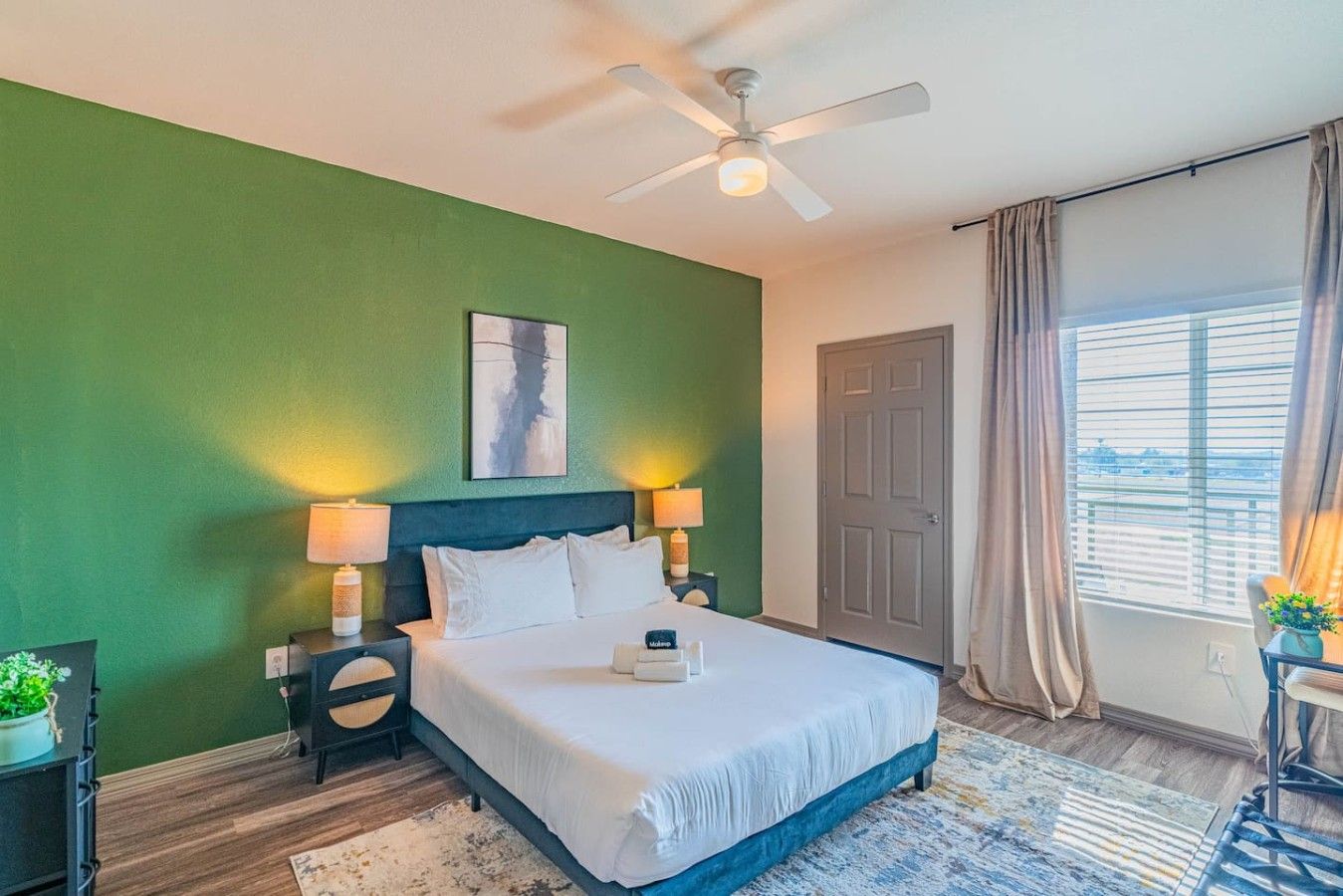 A hotel room with a bed, bedside lamps, a ceiling fan, a window with curtains, and a view of the outside.