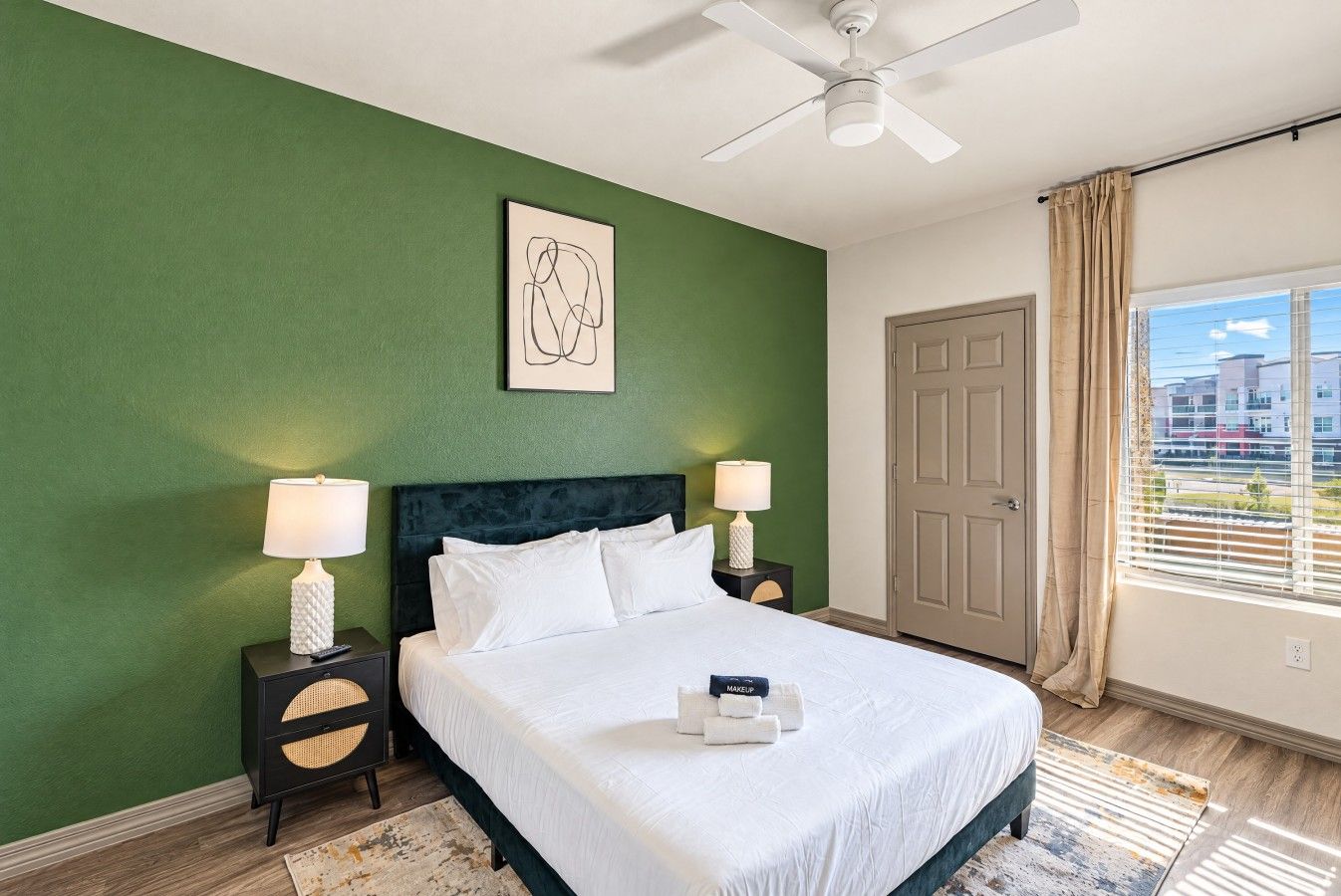 Une chambre d’hôtel comprenant un lit, deux tables de chevet avec des lampes, un ventilateur de plafond, une porte et une fenêtre avec des rideaux.
