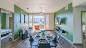 Un espace de vie compact comprenant un coin repas, un canapé, un téléviseur fixé au mur et une fenêtre avec des stores.