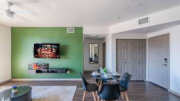 A modern living room with a green accent wall, a flat-screen TV, a dining table with chairs, and a ceiling fan.