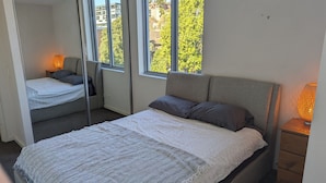 Een slaapkamer met een groot bed, een kleinere bank en een raam met uitzicht op gebouwen en bomen.