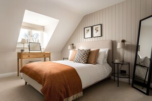 A bedroom with a sloped ceiling, a bed with a brown throw blanket, a bedside table with a lamp, a small round table with a cup, and a mirror. - Beautifully refurbished two bedroom - 1 Horseshoe Cottages (Kingham)