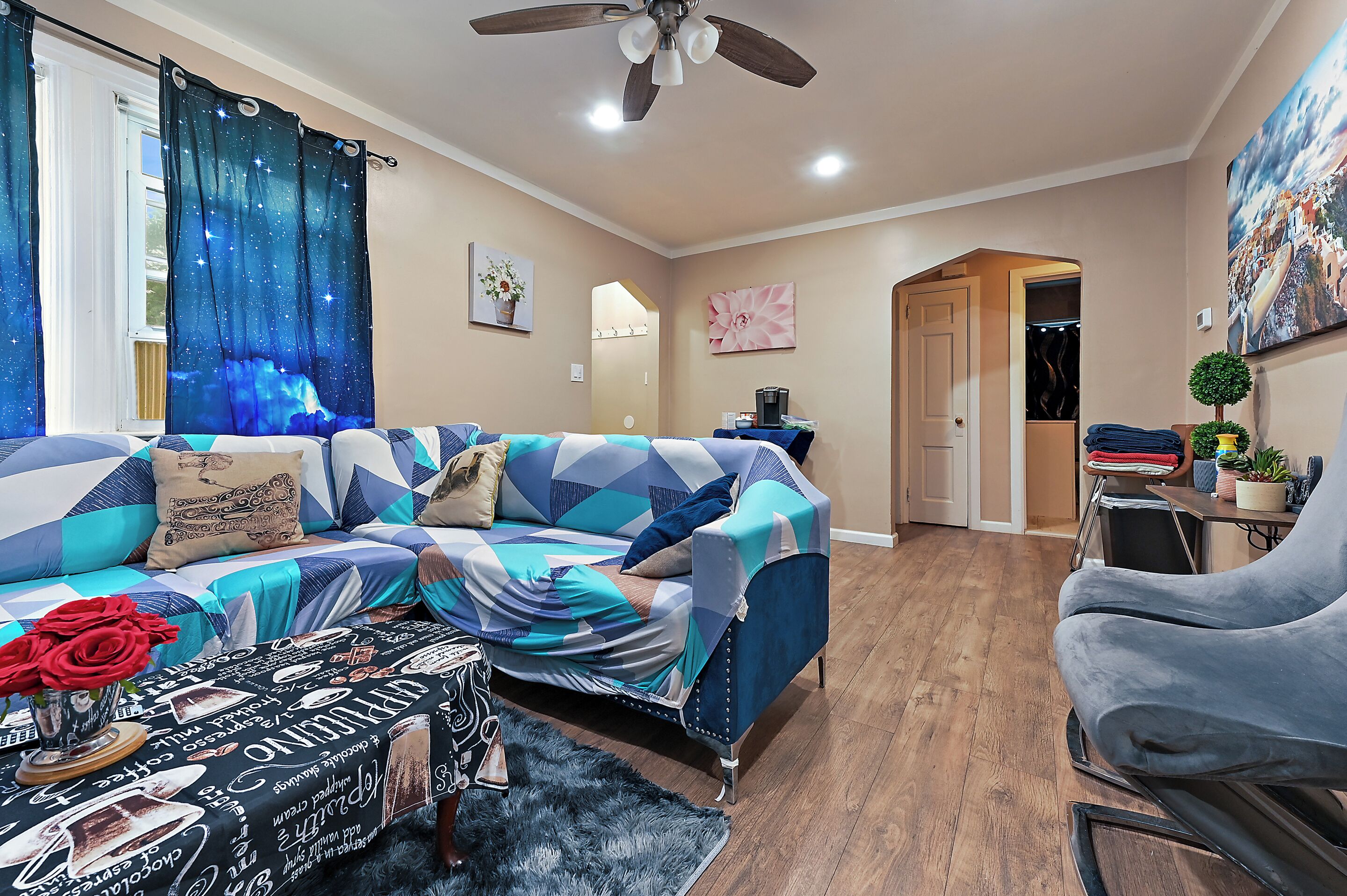 A living room with a sofa, a coffee table, and a chair. There is a ceiling fan and a door leading to another room.