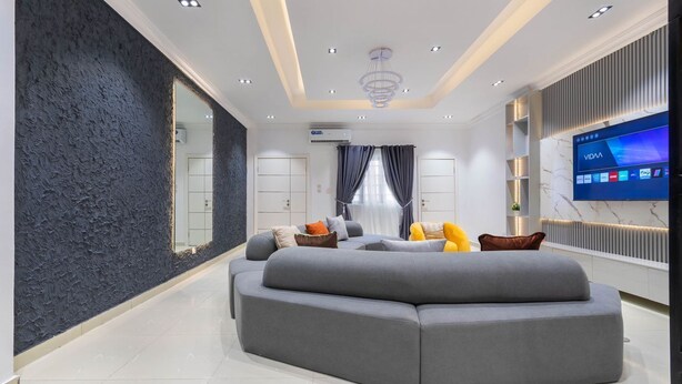Ein modernes Wohnzimmer mit einem grauen Ecksofa, einem großen Flachbildfernseher und einer dunkel strukturierten Akzentwand.