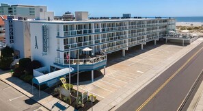 A multi-story hotel with a poolside area, a parking lot, and a view of the beach.