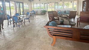 Un espacio de estar al aire libre cubierto con muebles y sillas de madera, ventanas amplias y un ventilador de techo.