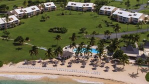 Vista aérea de un resort con varias construcciones, una playa con tumbonas, palmeras y una piscina.