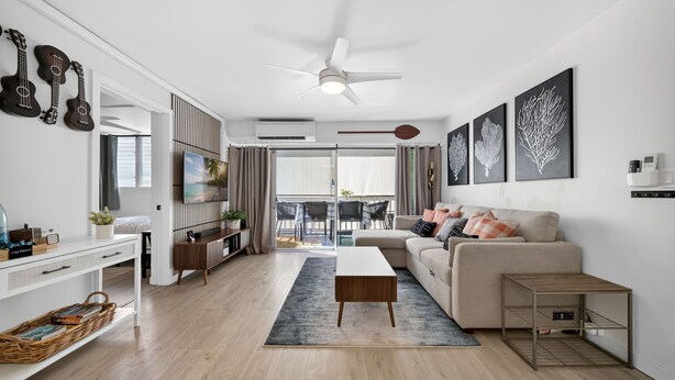 Una sala de estar moderna con sofá, mesa de centro y televisión. Hay un ventilador de techo, una zona de comedor con sillas y vistas al exterior.