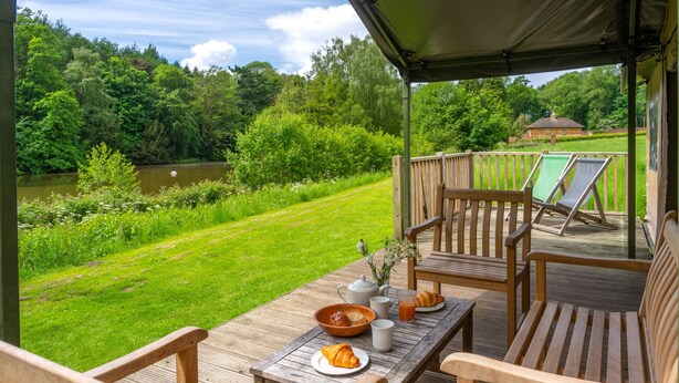 Une terrasse en bois avec des meubles d’extérieur, une table dressée pour le petit-déjeuner et une vue sur un lac et la verdure.
