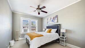 A bedroom with a bed, a ceiling fan, a radiator, a window with blinds, a nightstand with a lamp, and a painting on the wall. - Serene Suburban Stay: Family-Friendly w/EZ Parking (Berwyn)