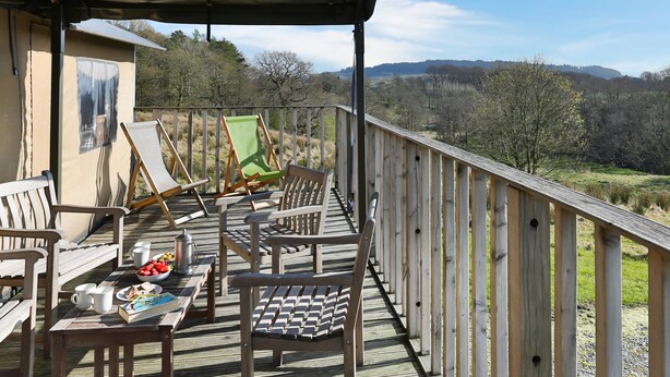 Eine Holzterrasse mit Sitzgelegenheiten, ein Tisch mit Speisen und Getränken und eine schöne Aussicht auf Bäume und Hügel.