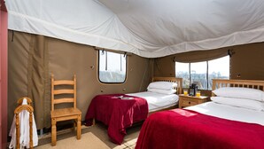 Ein Zeltraum mit zwei Betten, einem Holzstuhl, einem kleinen Tisch mit einer Lampe und einem Fenster.