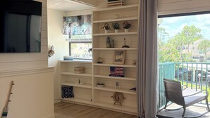 Una habitación con televisión, una estantería con diversos objetos, una mesita con un juego y vistas a un balcón con una silla.