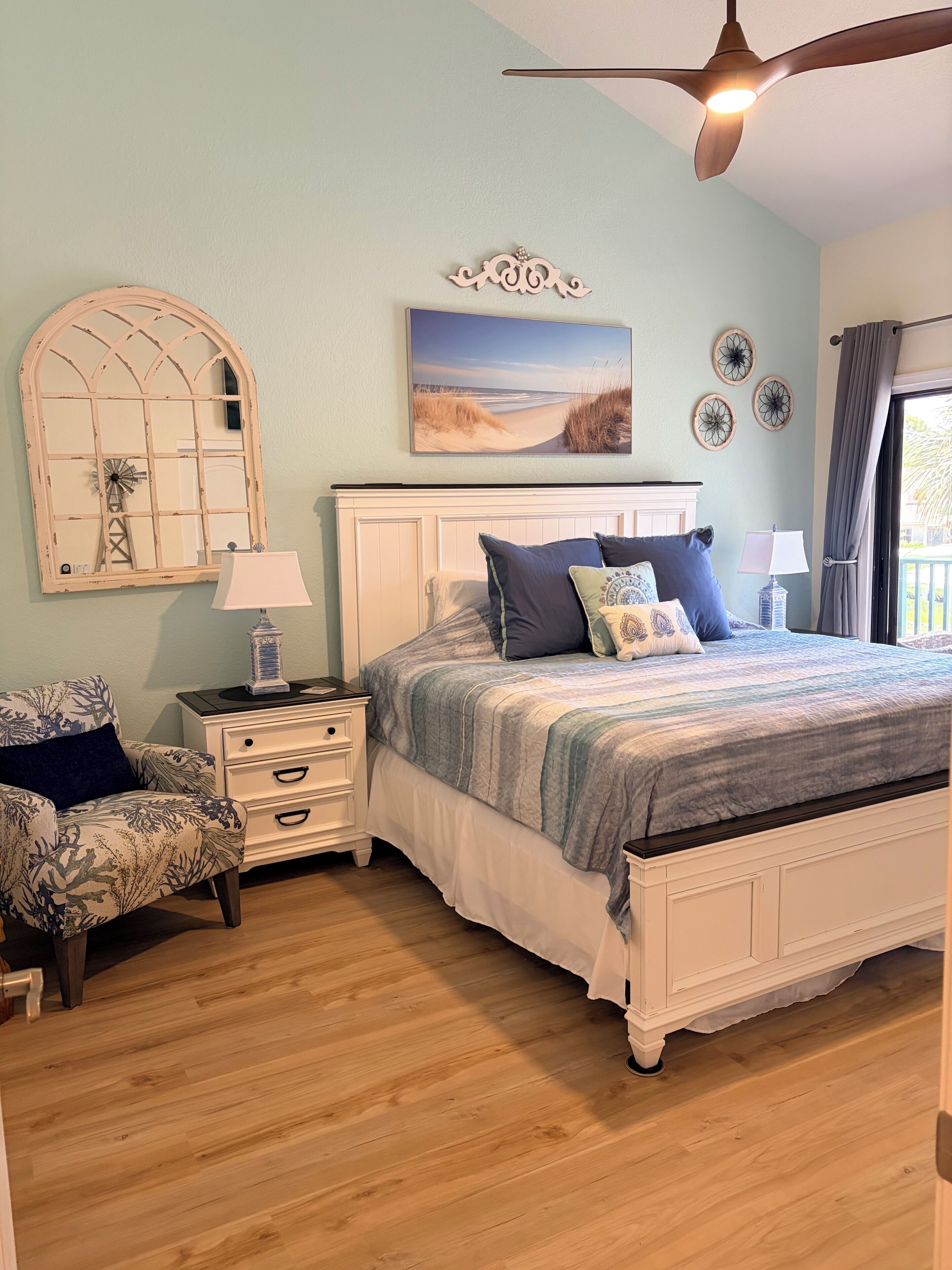 Een slaapkamer met een bed, nachtkastjes, een stoel, een raam met decoratieve lijst, een plafondventilator en wanddecoratie.