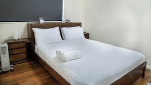 Ein ordentlich bezogenes Bett mit weißen Leinen und einem zusammengerollten Handtuch, ein Nachttisch mit Lampe, einer Uhr und einem kleinen dekorativen Gegenstand.