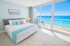 Een slaapkamer met een groot bed, uitzicht op de oceaan, een balkon met een tafel en stoelen, en een bord op de muur.