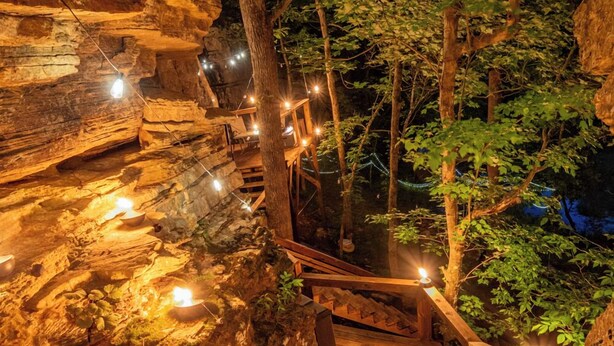 Una escalera de madera que desciende por un sendero rocoso rodeado de árboles, con luces a lo largo de los peldaños.