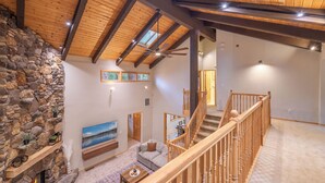 A spacious living area with a stone fireplace, wooden ceiling beams, a staircase, and a large window. - Spacious 5BR Carnelian Grand Retreat, Hot Tub, perfect for the whole family (Carnelian Bay)