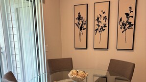 Un comedor con mesa de vidrio rodeada por cuatro sillas tapizadas, un florero con flores y tres láminas botánicas enmarcadas en la pared.