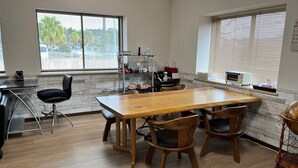 Un coin repas avec une table et des chaises en bois, un espace cuisine équipé d’une cuisinière et d’un évier, et de grandes fenêtres avec des stores.